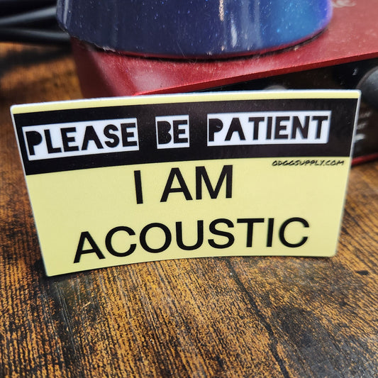 Sticker on a guitar neck with text 'Please be patient, I am acoustic' on a wooden surface.
