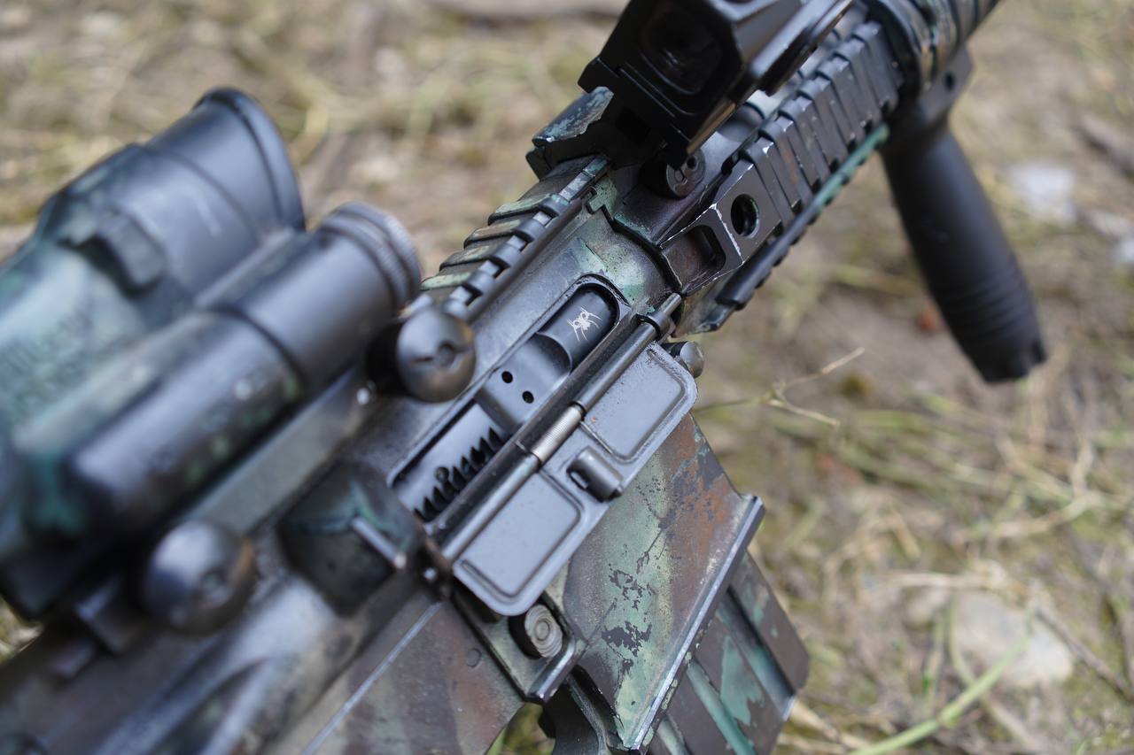 Close-up of a tactical rifle with a green and black camouflage finish, mounted scope, and black handguard, photographed outdoors on a blurred grassy ground.