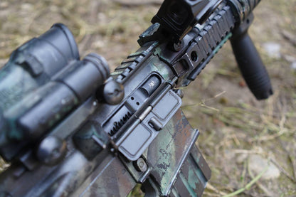 Close-up of a tactical rifle with a green and black camouflage finish, mounted scope, and black handguard, photographed outdoors on a blurred grassy ground.