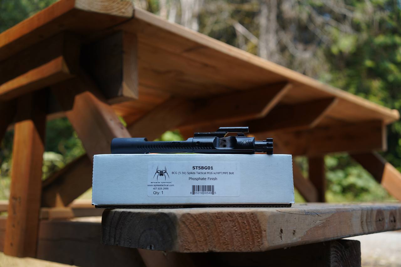 A black rifle scope mounted on top of a white box labeled STS86G01, placed on a wooden picnic table with an outdoor forest background.